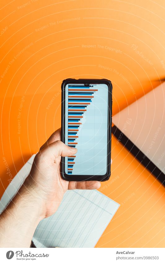Eine Hand hält ein Telefon mit Statistiken über einen orangefarbenen Hintergrund mit Laptop und Notebook mit Finanzkonzept finanziell blanko Business Büro