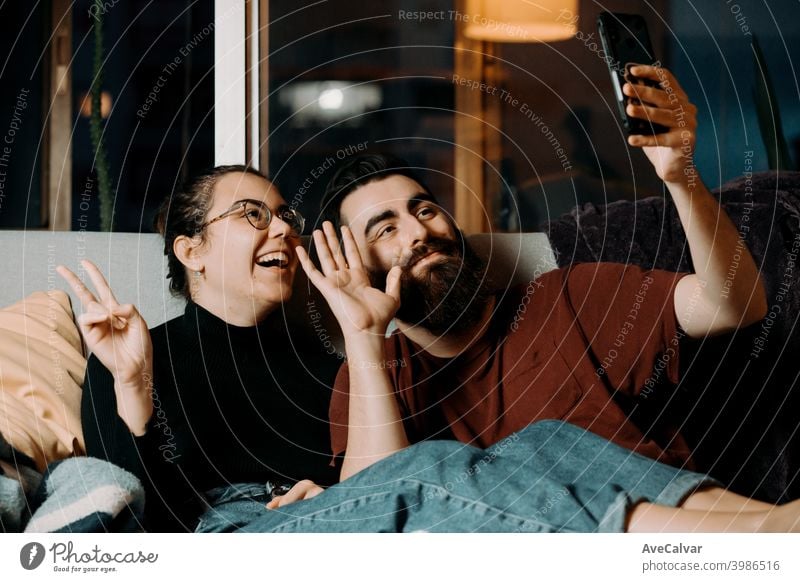 Ein junges Paar auf der Couch grüßt zum Handy und lächelt, während auf Pandemie anhänglich umarmend Humor Ehefrau Ehemann Foto Ethnizität lebend Partnerschaft
