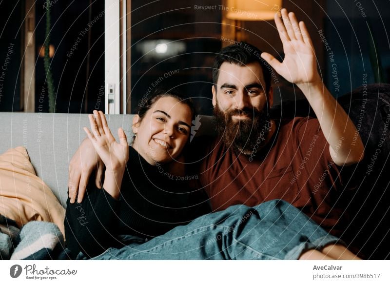 Ein junges Paar auf der Couch, das in die Kamera grüßt und lächelt, während es auf Pandemie anhänglich umarmend Humor Ehefrau Ehemann Foto Ethnizität lebend