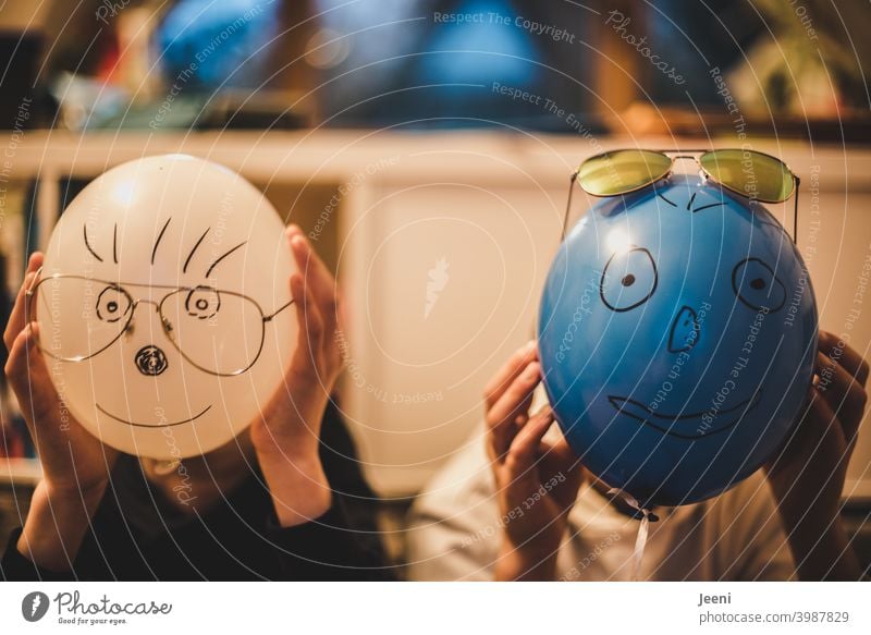 Mit lustigen Gesichtern bemalte Luftballons vor den Köpfen zweier Kinder | Aufgemalte Augen, Nase, Mund, Haare und aufgesetzte Brille | Hände halten den Luftballon fest