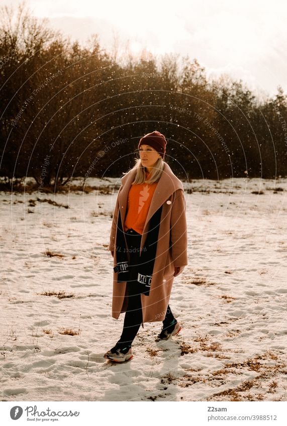 Modisch gekleidete Junge Frau geht bei leichtem Schneefall spazieren schneien winterwonderland frau junge frau mantel spaziergang schnee sonne licht natur
