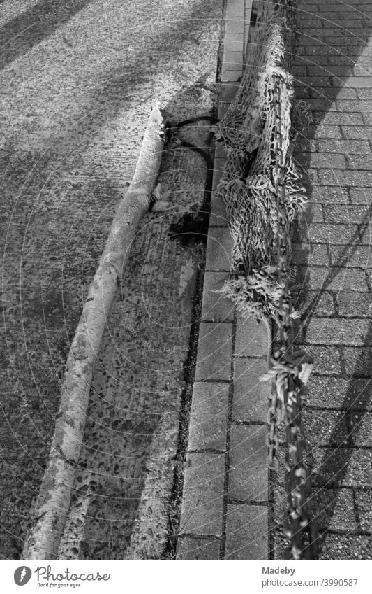 Über eine alte rostige Eisenkette hängende Fischernetze im Hafen von Neuharlingersiel an der Nordseeküste bei Esens im Landkreis Wittmund in Ostfriesland in Niedersachsen, fotografiert in klassischem Schwarz-Weiß