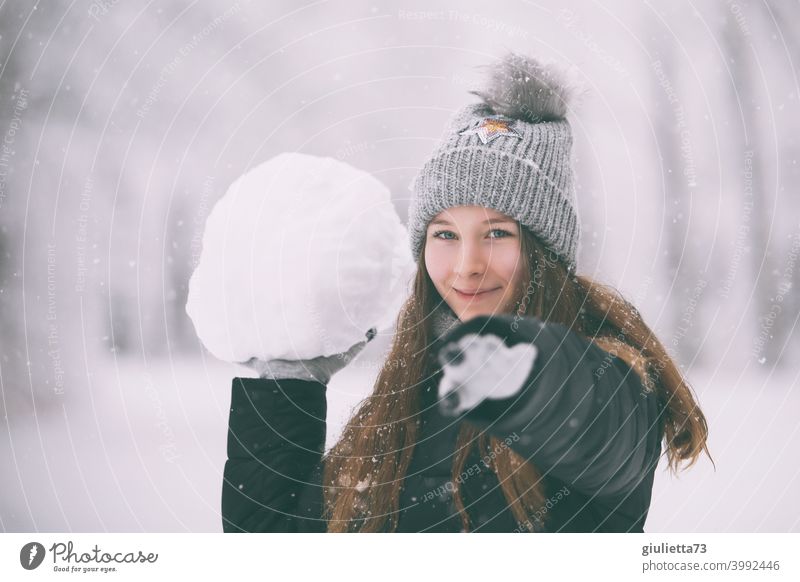 Glückliches, freches Teenager Mädchen im Winter mit riesengroßem Schneeball weiß Bommelmütze feminin Gedeckte Farben Schneeflocken verschneit langhaarig