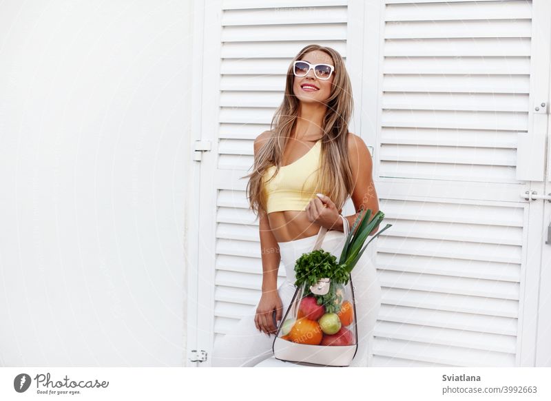 Ein junges Mädchen mit Sonnenbrille und Sportkleidung steht im Hintergrund der Tür mit einer Tüte voller Gemüse und Obst. Das Konzept der gesunden Ernährung und des gesunden Lebensstils