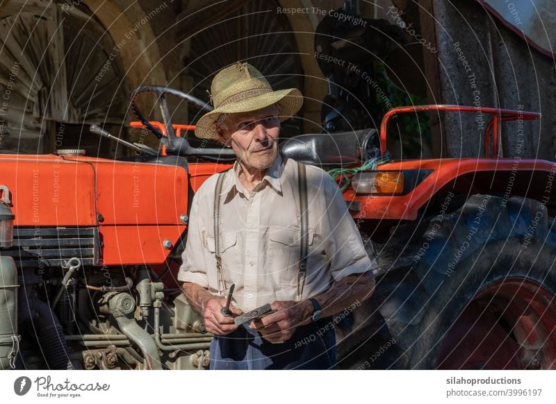 Alter Bauer arbeitet am roten Traktor unter dem Dach des Bauernhauses im Sonnenschein. Erwachsener gealtert Ackerbau Auto Automobil PKW Kaukasier Schriftzeichen