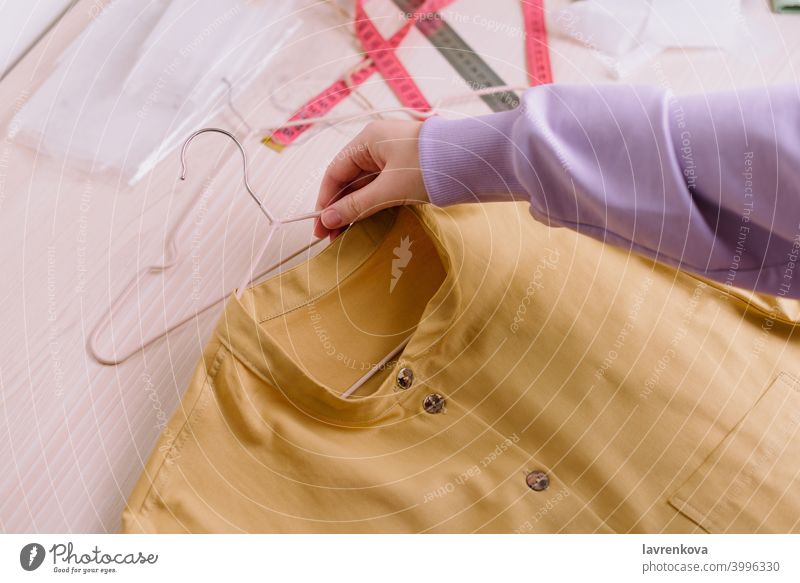 Draufsicht auf die Hände einer Näherin, die ein fertiges gelbes Hemd auf einem Bügel hält Werkstatt Job Kleiderbügel Unternehmer Frau Mädchen Schneider Nähen