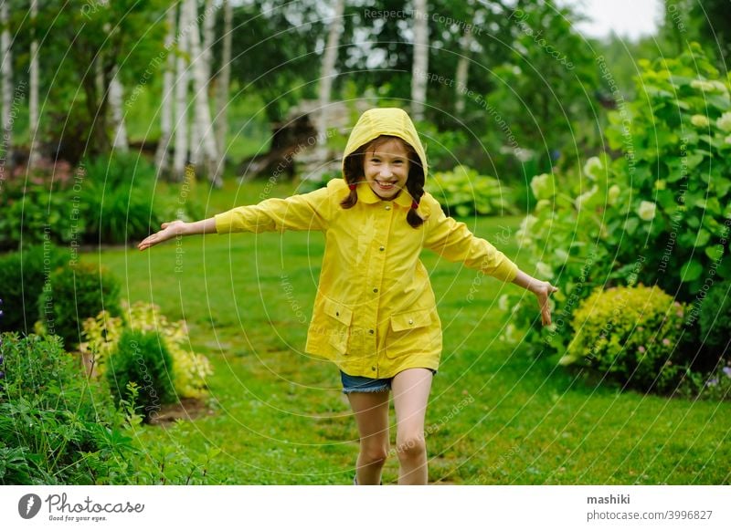 glückliches Kind Mädchen in gelben Regenmantel spielen und Spaß haben im Sommer Garten unter dem regen im Freien Wetter Stiefel regnerisch wasserdicht nass