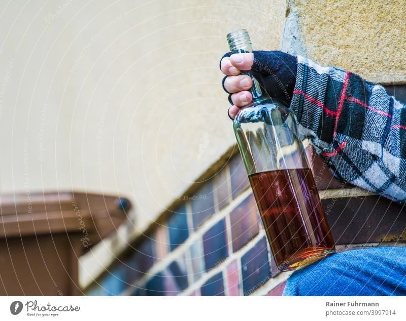 Nahaufnahme von einem anonymen Mann der an einem Gebäude sitzt und eine offene Schapsflasche in der Hand hält Alkohol Alkoholismus Sucht Mülltonne Säufer