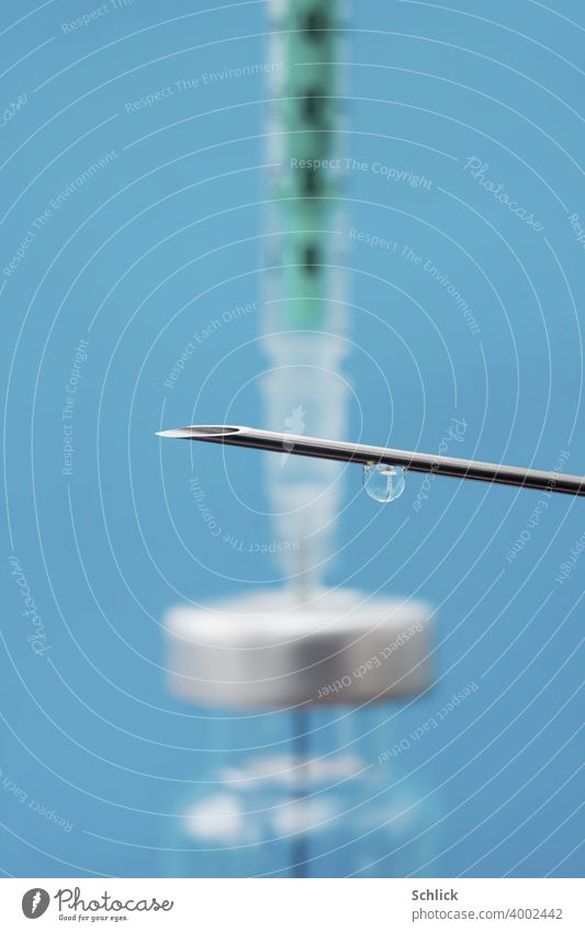 Ein Tropfen eines Impfstoffes hängt an einer Kanüle und zeigt das verkleinerte Abbild einer kleinen Glasflasche mit Septum und Spritze Makro mit geringer Tiefenschärfe und blauem Hintergrund