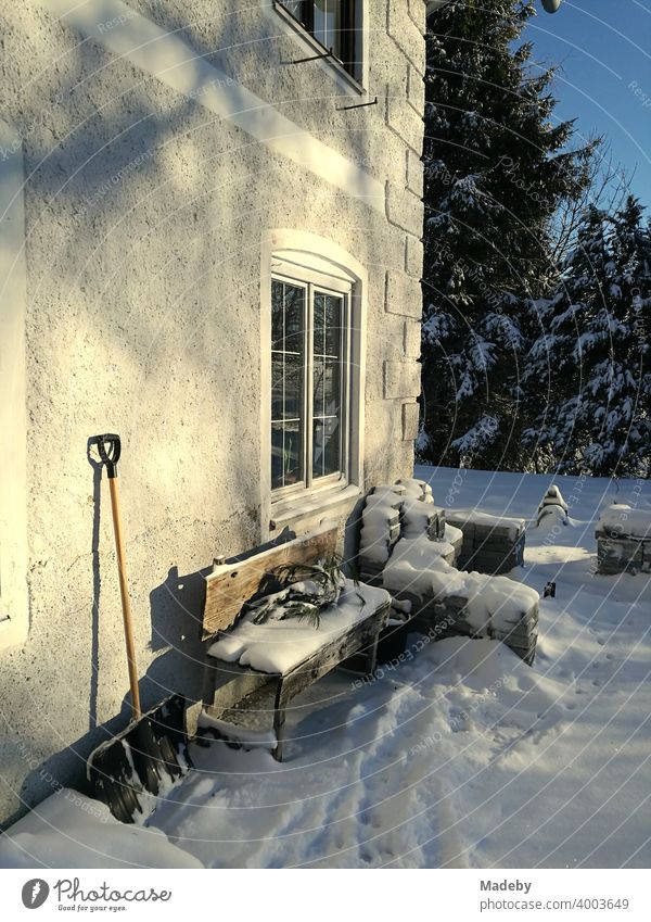 Verschneite Holzbank und angelehnte Schneeschaufel an einem alten Bauernhaus im Winter bei Sonnenschein in romantischer Schneelandschaft in Rudersau bei Rottenbuch im Kreis Weilheim-Schongau in Oberbayern