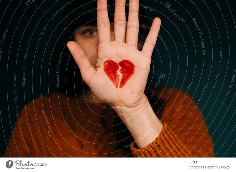 Eine Frau hält die Hand, auf der ein zerbrochenes rotes Herz gemalt ist, vors Gesicht. Konzept Liebeskummer. Beziehungsende Gefühle Partnerschaft Probleme