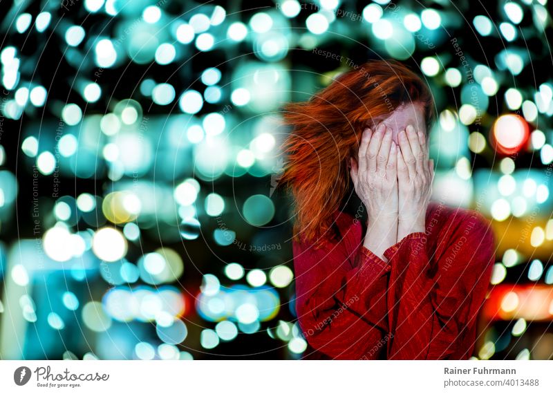 Eine rothaarige Frau hält sich die Hände vor das Gesicht. Im Hintergrund leuchtet das Bokeh heller Lichter. Probleme Kopfschmerzen abstrakt Unschärfe