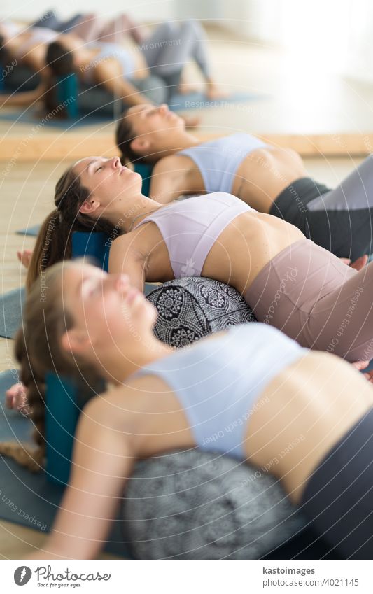 Restorative Yoga mit einem Bolster. Gruppe von drei jungen, sportlichen, attraktiven Frauen im Yogastudio, liegend auf einem Bolster-Kissen, dehnend und entspannend während Restorative Yoga. Gesunder aktiver Lebensstil