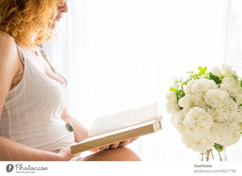 Schwangere Frau, die ihren Bauch berührt, während sie ein Buch liest schwanger Bett Schwangerschaft lesen Rotschopf rote Haare Morgen jung Mädchen Fenster