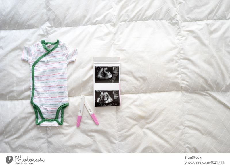 Schwangerschaftstests, Babykleidung und Ultraschalluntersuchung auf dem Bett, Ansicht von oben Prüfung positiv Zubehör Scan weiß Hintergrund Draufsicht