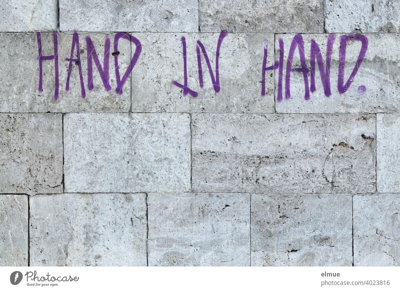 "HAND IN HAND" steht in lila Druckbuchstaben an der grauen Natursteinwand / gemeinsam Hand in Hand Wand Fassade Losung Graffito Graffiti Schmiererei