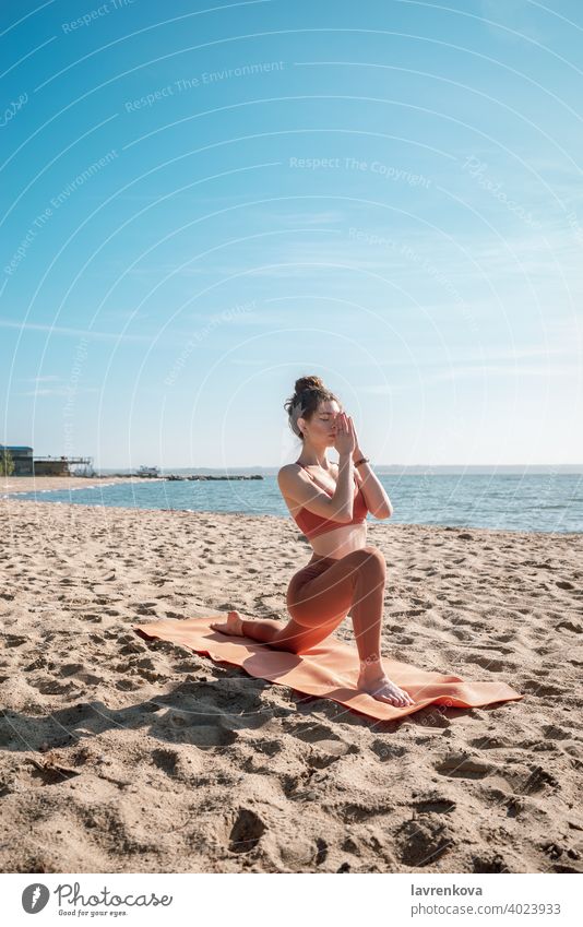 Junge Frau in orangefarbenem Oberteil und Leggins beim Üben der Sichel-Lunge-Pose (Anjaneyasana) am Strand am Morgen Übung Yoga jung praktizieren im Freien
