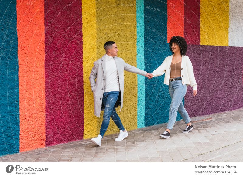 Junges glückliches Paar, das sich an den Händen hält und durch einen Bürgersteig gegen eine bunte Wand läuft Straßenbelag farbenfroh streifen jung Beteiligung
