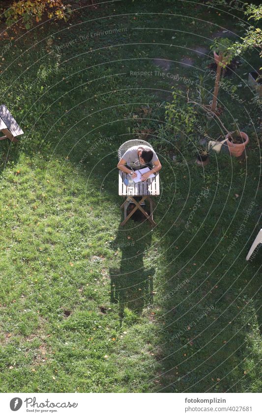 Vogelperspektive auf einen jungen Mann, der im Garten an einem kleinenTisch sitzt  und in ein Heft schreibt junger Mann Gartentisch lernen studieren schreiben