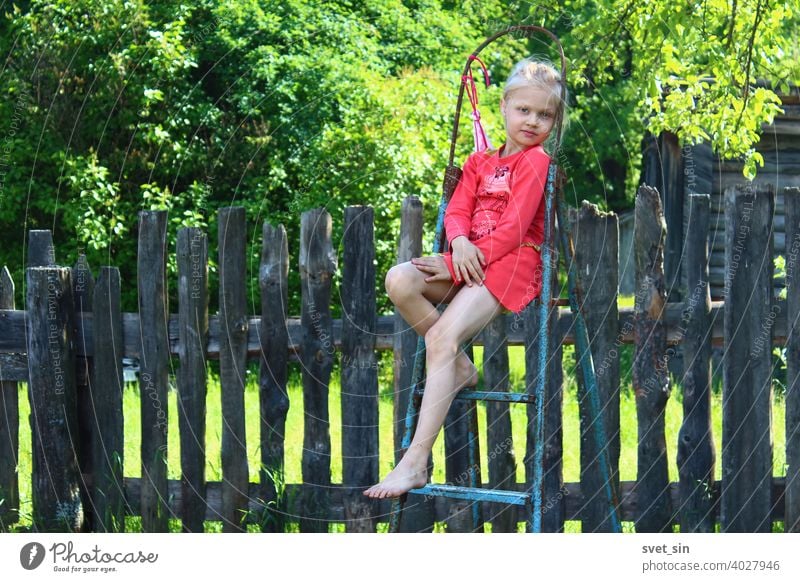 Barfuß Sommer auf dem Dorf. Porträt eines kleinen Mädchens mit blonden Haaren in einem roten Kleid, das auf einer Trittleiter auf dem Lande vor dem Hintergrund eines alten Holzzauns und grünem Laub an einem sonnigen Sommertag sitzt.