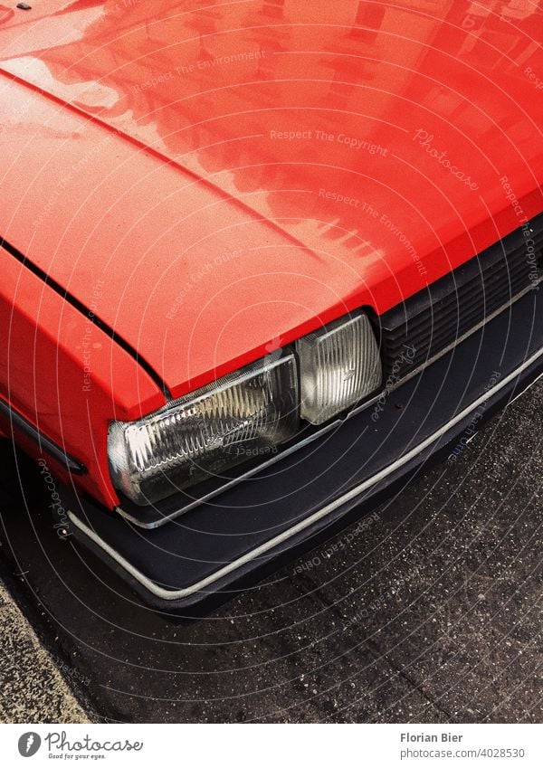 Rot lackierte Motorhaube und eckiger Scheinwerfer sowie Kühlergrill und Kunstoffstoßstange eines geparkten Autos auf der Straße rot transparent Stoßstange