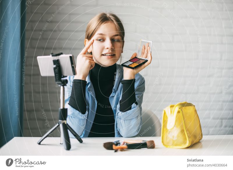 Ein Teenager-Mädchen schminkt sich und zeigt, wie man Rouge aufträgt, während sie mit ihrem Smartphone auf einem Stativ zu Hause ein Video für ihren Blog aufnimmt