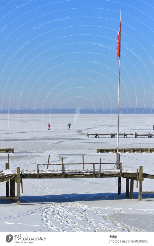 zugefrorener See mit Schnee, Spuren, Holzstegen, einer Fahnenstange und im Hintergrund zwei Menschen, die Schlittschuh laufen Winter Frost Eis Kälte Steg