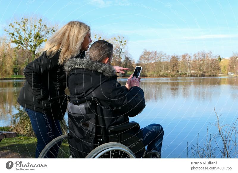 Konzept einer Person mit einer körperlichen Behinderung. Ein Mann in einem Rollstuhl mit einer Frau, die neben ihm steht. Paar, das Technologie verwendet, während es auf ein Smartphone schaut. Ländliche Szene an einem See.