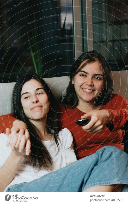 Ein schönes Paar von zwei Frauen, die den Tv in einer modernen Wohnung auf der Couch schauen, während lächelnd anhänglich Antwort an Kuscheln umarmend Freunde