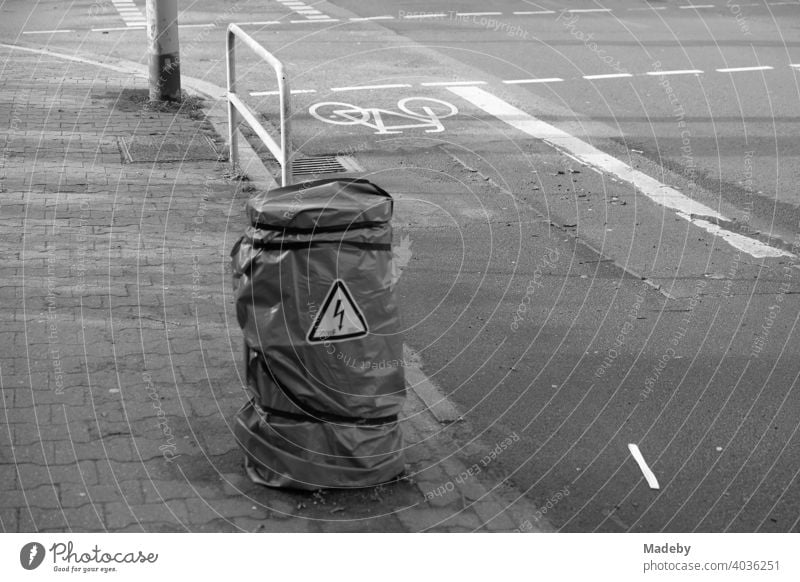 Warnung vor Hochspannung an einem mit Folie umwickelten Pfeiler zwischen Bürgersteig und Radweg im Westend von Frankfurt am Main in Hessen, fotografiert in neorealistischem Schwarzweiß