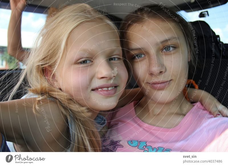 Lächelnde Schwestern auf einer Autofahrt: blond und braunhaarig. Zwei Mädchen unterschiedlichen Alters lächeln, umarmen sich, im Inneren des Autos. Teenager