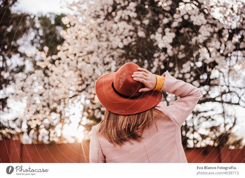 Rückenansicht Porträt der jungen Frau trägt modernen Hut bei Sonnenuntergang im Park. schönen Mandelbaum Blumen Hintergrund. Frühling und Blüte Bäume im Freien