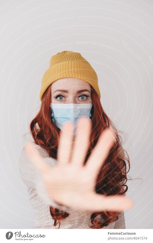 Junge rothaarige Frau mit gelber Hipster Beanie Mütze , medizinischer Schutzmaske Maske und ausgestreckter Hand Richtung Kamera als Zeichen für bitte Abstand halten