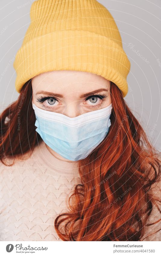Junge rothaarige Frau mit gelber Hipster Beanie Mütze und medizinischer Schutzmaske Maske schaut mit eindringlichem Blick in Kamera (Portrait)