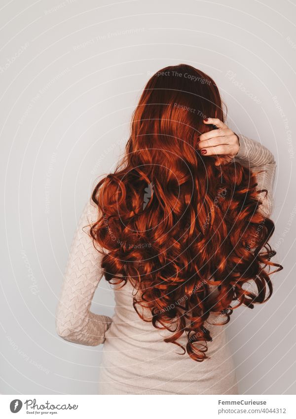 Rückansicht einer Frau mit langen roten lockigen Haaren in hautfarbenem Pullover die sich mit einer Hand ins Haar fasst Neutraler Hintergrund Textfreiraum oben