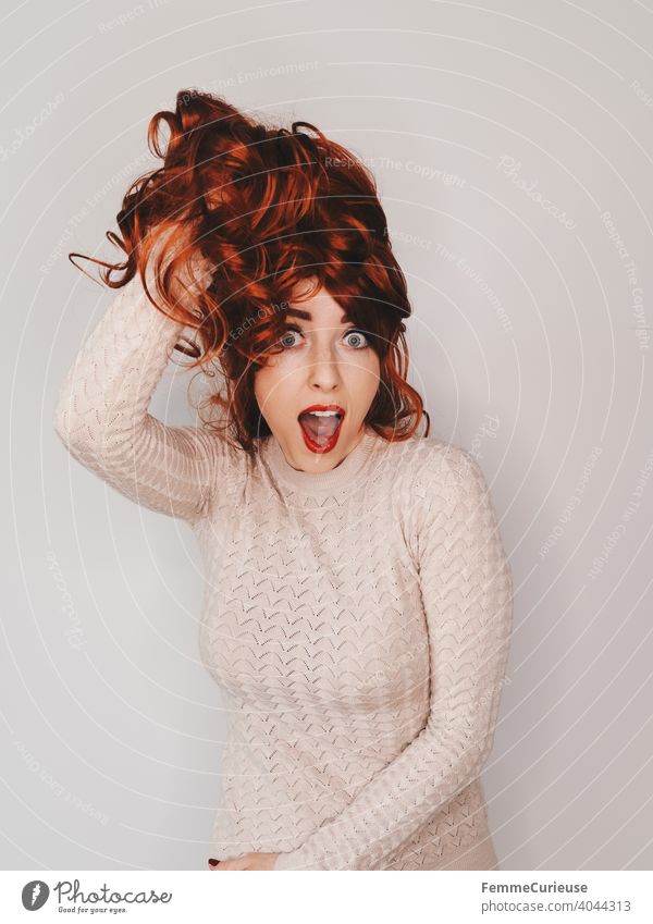 Frau hält ihre langen lockigen roten Haare mit einer Hand nach oben und posiert mit geöffentem Mund Neutraler Hintergrund Textfreiraum oben Textfreiraum links
