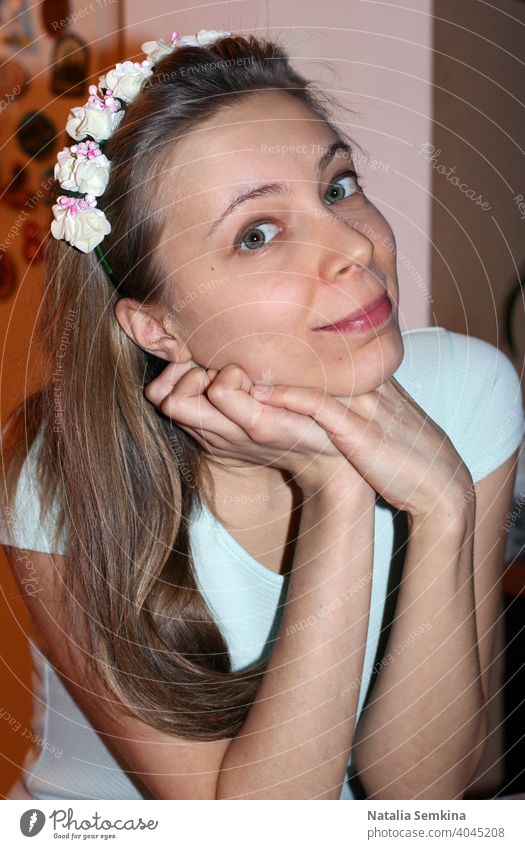 Attraktives junges Mädchen in hellem Kleid und Blumenkranz auf dem Kopf, das mit auf die Hände gestütztem Kopf sitzt und die Kamera in Nahaufnahme betrachtet. Vertikale Ausrichtung.