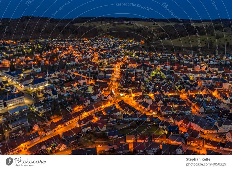 Luftaufnahme über eine beleuchtete Stadt mit Autos an den Straßen und orangefarbenen Lichtern von Laternen und einem Hügel hinter der kleinen Stadt. Nacht
