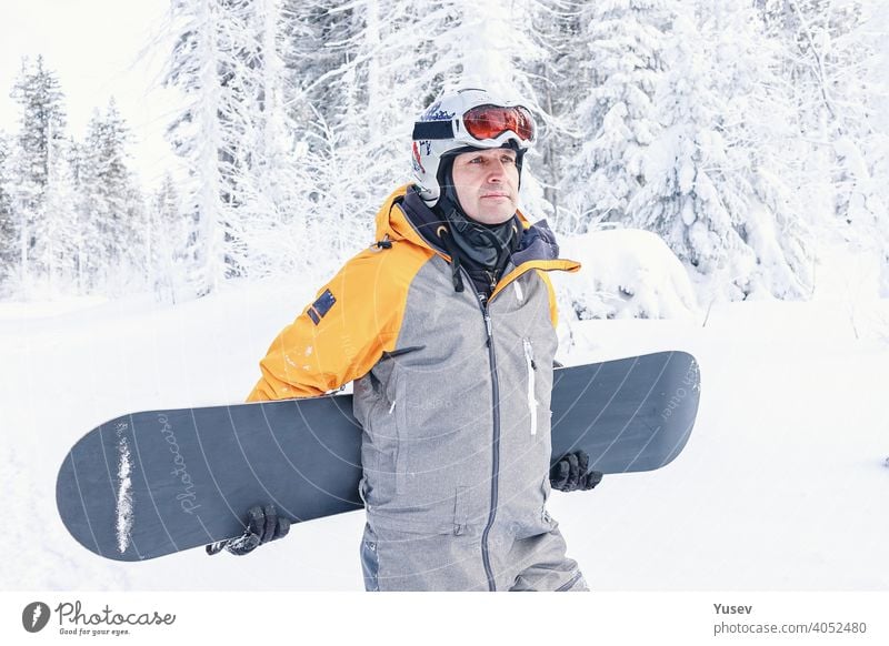 Schöne kaukasische Mann in einem hellen gelben und grauen Overall, weißen Helm und Brille hält sein Snowboard. Wintersport, Freizeitbeschäftigung. Urlaub in den Bergen. Vorderansicht.