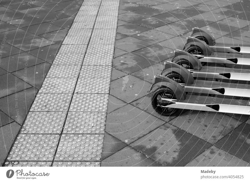 E-Roller aufgereiht zur Vermietung neben einem taktilen Leitsystem für Sehbehinderte auf grauem Straßenpflaster in der Innenstadt von Frankfurt am Main in Hessen, fotografiert in neorealistischem Schwarzweiß