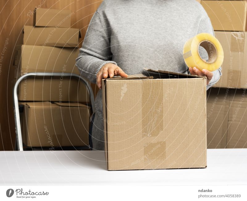 Frau im grauen Pullover hält eine Rolle Klebeband und packt braune Kartons auf einem weißen Tisch, hinter einem Stapel von Kartons Lifestyle wirklich viele