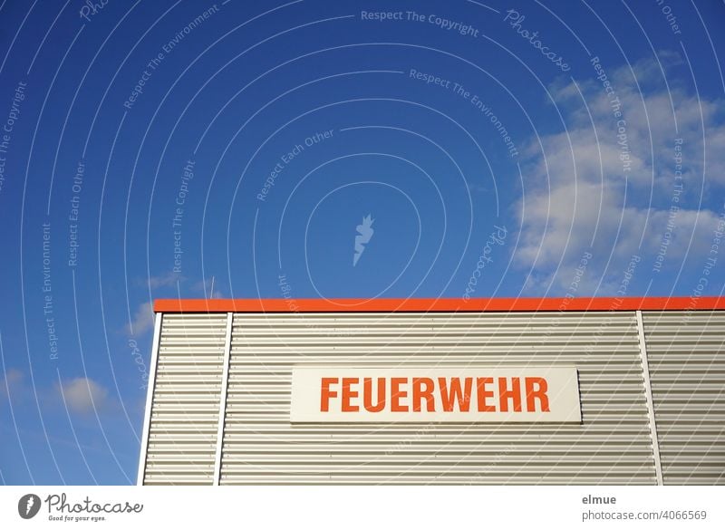 Teilansicht eines Zweckgebäudes mit Metallverkleidung und der Aufschrift in rot "FEUERWEHR" vor blauem Himmel mit Schönwetterwolken / Rettungsdienst / Notruf 112