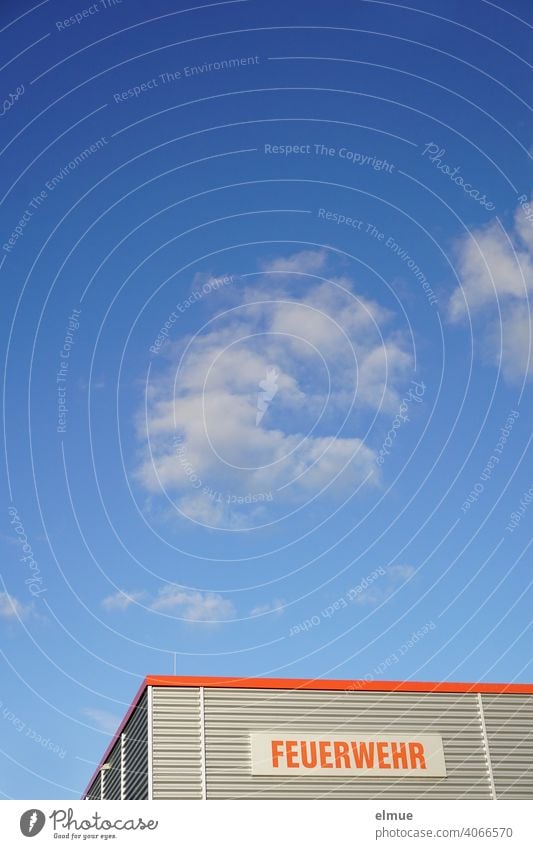 Zweckgebäude mit grauer Wellblechverkleidung und einem Schild mit roter Aufschrift "FEUERWEHR"  vor blauem Himmel mit Schönwetterwolken / 112 / Rettungsdienst