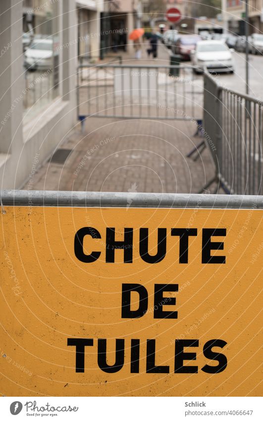 Vor einer Absperrung steht auf einem gelben Schild in französischer Sprache CHUTE DE TUILES Dachdeckerarbeiten oder Sturmschaden Chute de Tuiles Gehweg