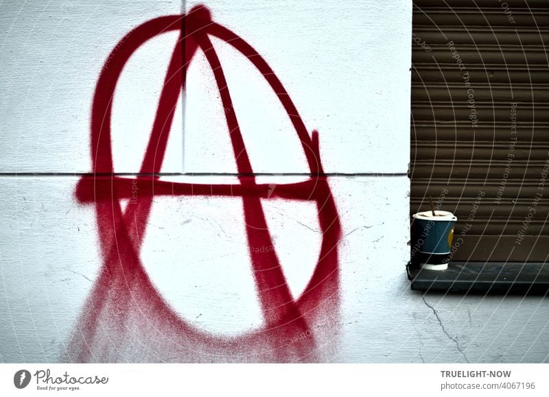 Ein Sprayer hat auf eine weisse Hauswand groß und in roter Farbe das bekannte Signet der Anarchisten oder Anarchos gesprüht: großes A im Kreis. Der  Kaffeebecher auf der Fensterbank daneben vor geschlossenem braunem Rolladen wirkt passend