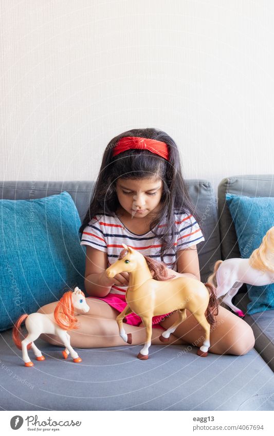 Kleines Mädchen spielt mit ihren Spielzeugpferden auf dem Sofa allein Appartement Schwarzes Haar sorgenfrei Freizeitkleidung Kind Kindheit Farbbild bequem