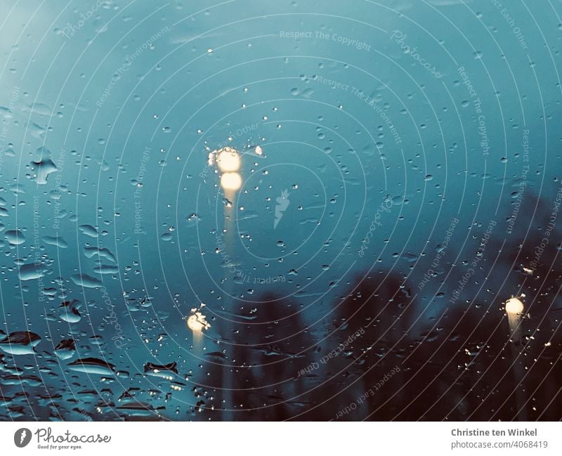 Blick durch ein nasses Fenster auf die Abenddämmerung zur blauen Stunde. Regenwetter mit verschwommenen Bäumen und Straßenlaternen verregnet Dämmerung