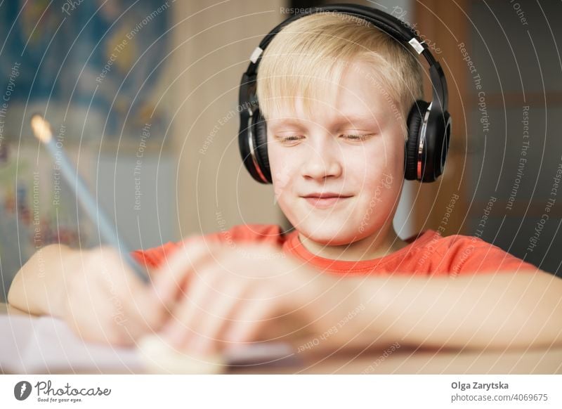 Junge mit Kopfhörer schreibt und lächelt. Lächeln schreibend Kind im Innenbereich niedlich Hausaufgabe Spaß Kaukasier blond Hand Bleistift Behaarung Gesicht