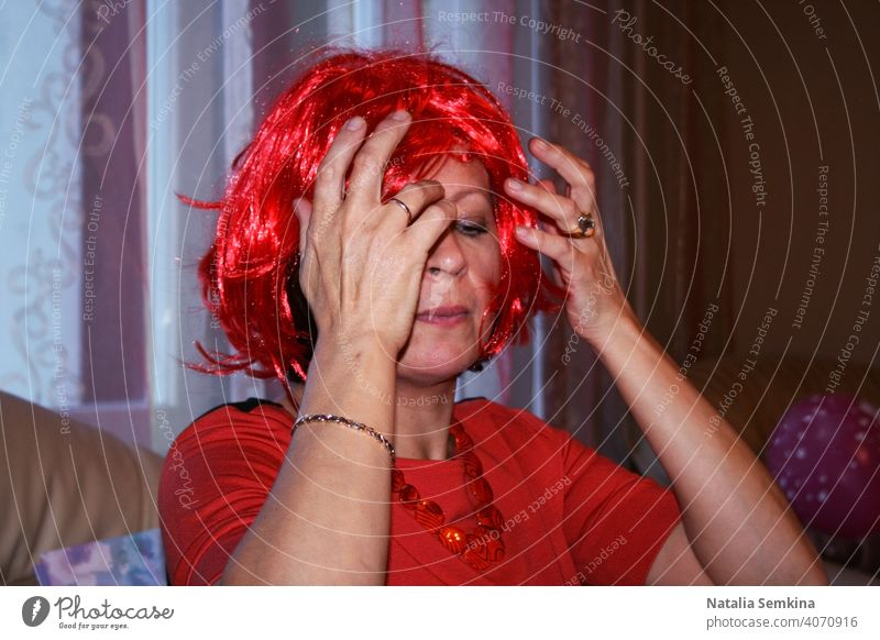 Eine Frau sitzt auf dem Sofa und passt feuerrote Perücke auf ihrem Kopf auf einer Geburtstagsfeier an. Kopf und Schultern Porträt. Party zu Hause.