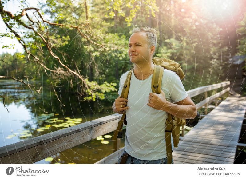Reifer Mann erkundet die finnische Natur im Sommer, geht über die Brücke. Wanderer mit großen Rucksack Reisen in Wäldern. Sommer skandinavischen Landschaft von Seen und Wäldern.